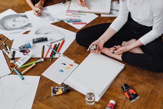 Artists sitting on wooden floor creating art with pencils and paints, showcasing creativity and design process.